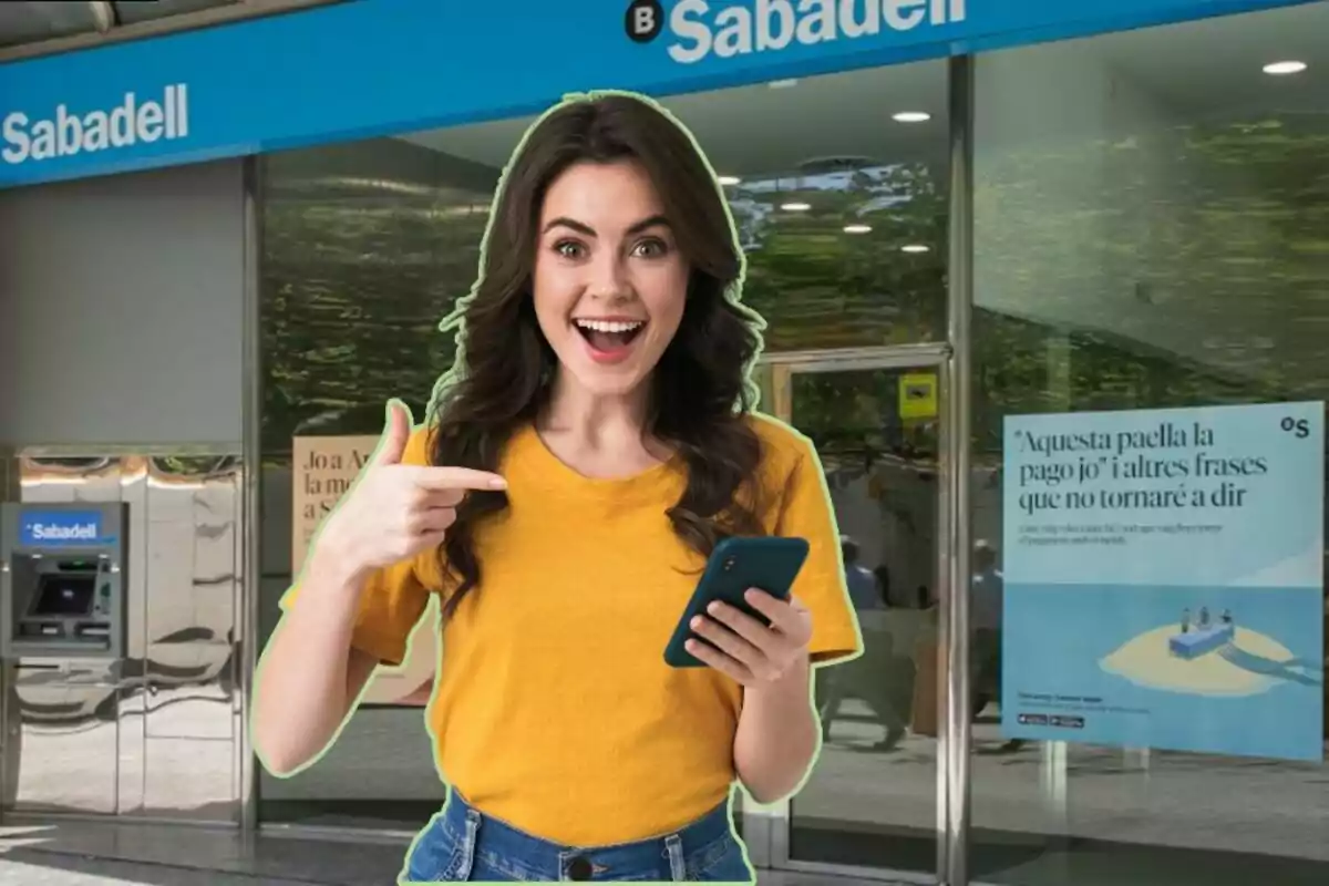 Una mujer sonriente con una camiseta amarilla sostiene un teléfono móvil y señala hacia él, mientras está de pie frente a una sucursal del banco Sabadell.