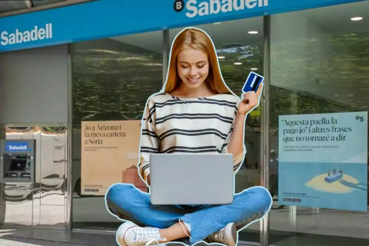 Una joven sonriente con una tarjeta de crédito y una computadora portátil frente a una sucursal del banco Sabadell.