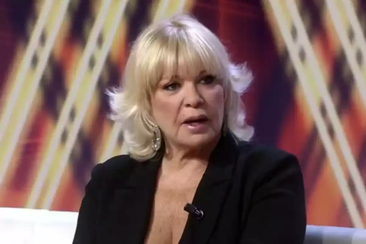 Bárbara Rey usando un saco negro y aretes grandes, sentada en un set de televisión con fondo colorido. Bárbara Rey usando un saco negro y aretes grandes, sentada en un set de televisión con fondo colorido.