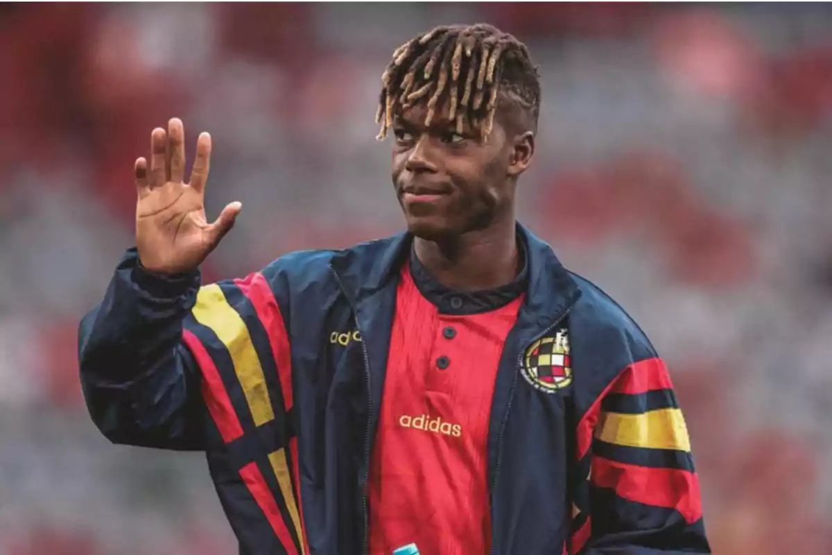 Un jugador de fútbol con una chaqueta azul con detalles en rojo, amarillo y negro, y una camiseta roja, saluda con la mano levantada. Un jugador de fútbol con una chaqueta azul con detalles en rojo, amarillo y negro, y una camiseta roja, saluda con la mano levantada.