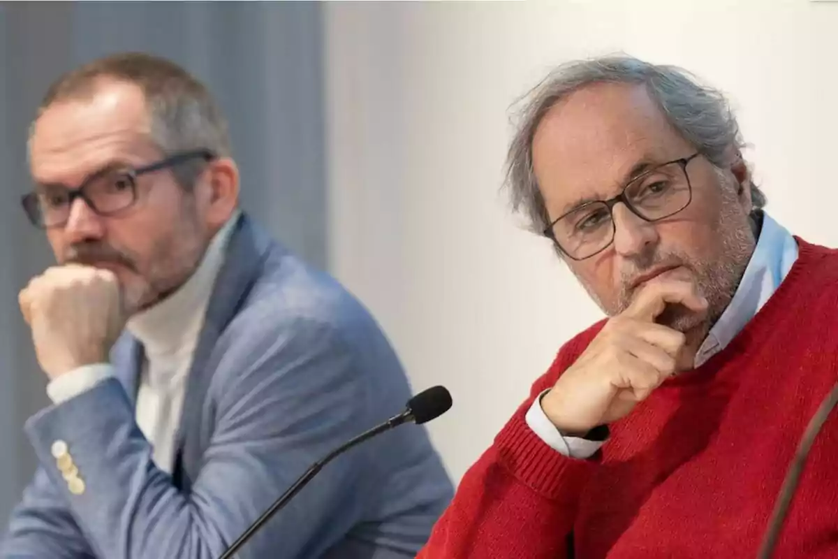 Dos hombres sentados en una mesa durante una conferencia, ambos con expresión pensativa y usando gafas.