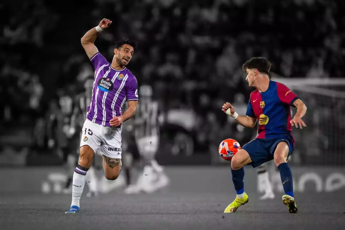 Dos jugadores de fútbol en acción durante un partido uno con uniforme morado y blanco y el otro con uniforme azul y rojo en un estadio. Dos jugadores de fútbol en acción durante un partido uno con uniforme morado y blanco y el otro con uniforme azul y rojo en un estadio.