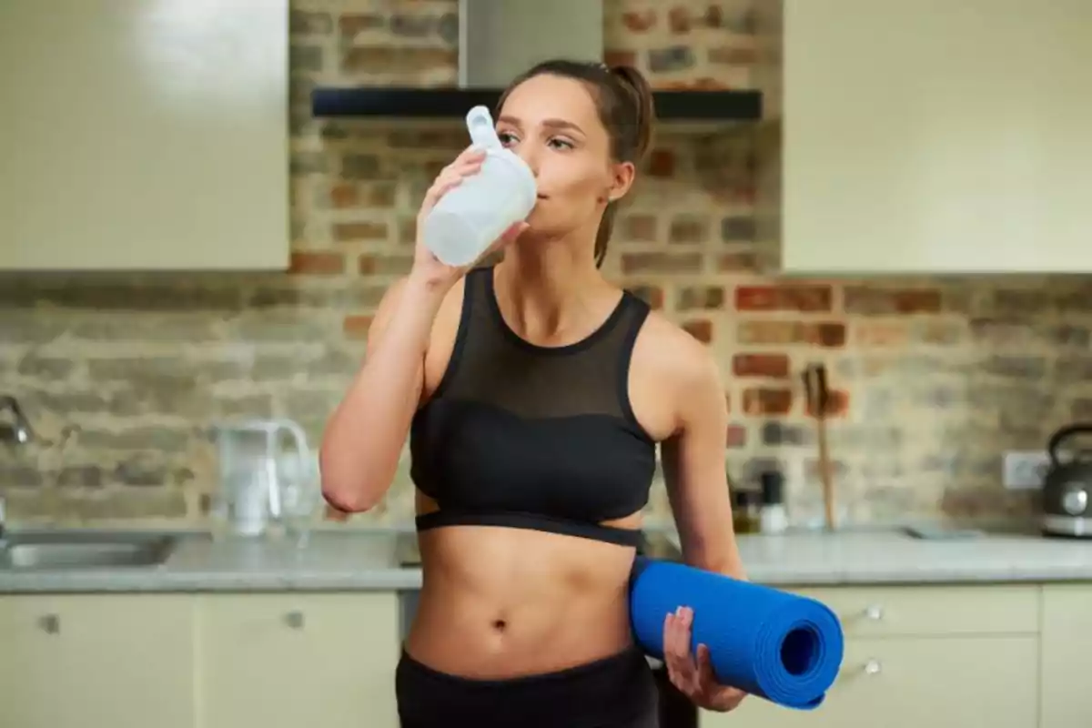 Mujer en ropa deportiva bebiendo de una botella mientras sostiene una esterilla de yoga en una cocina.