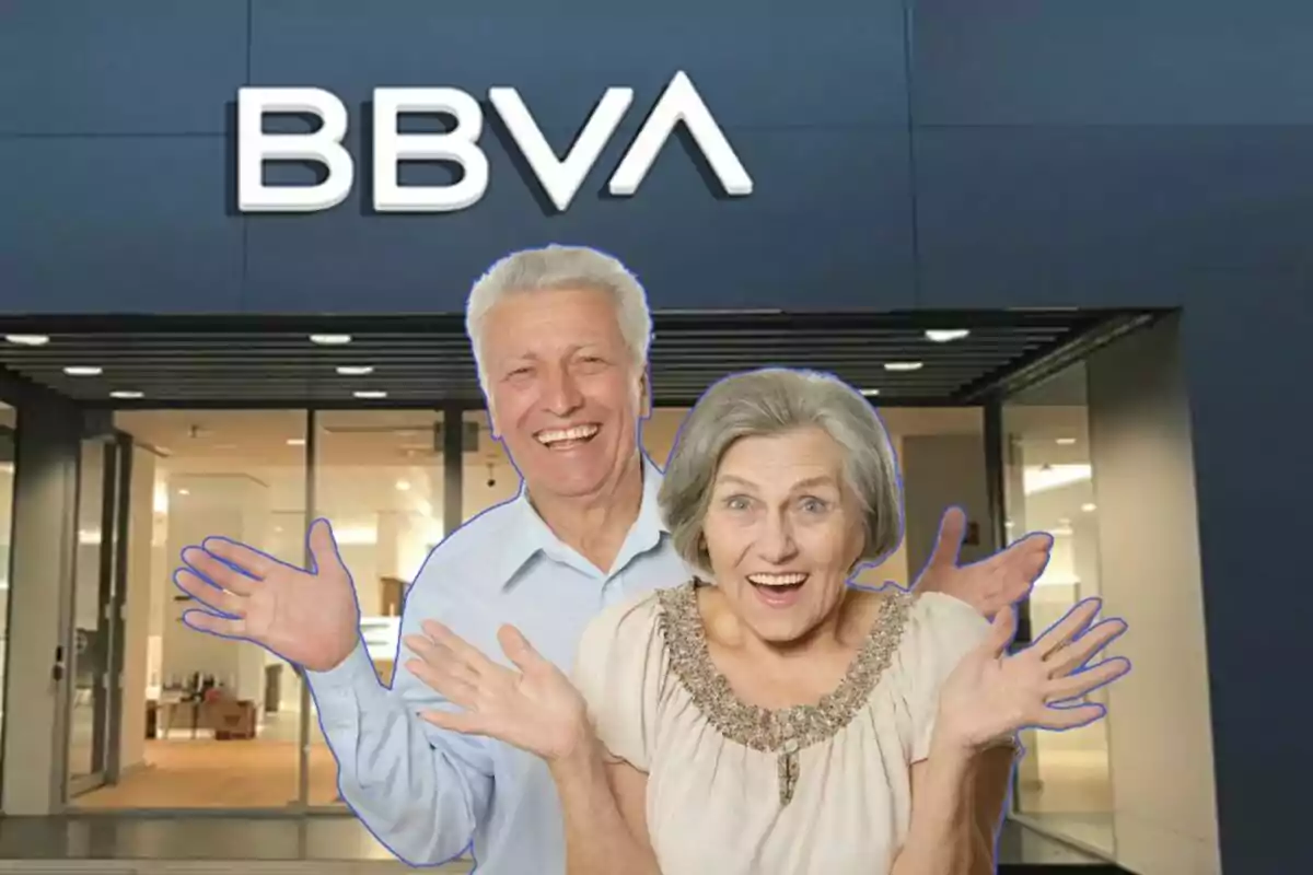 Una pareja de personas mayores sonríe y levanta las manos frente a una sucursal del banco BBVA.