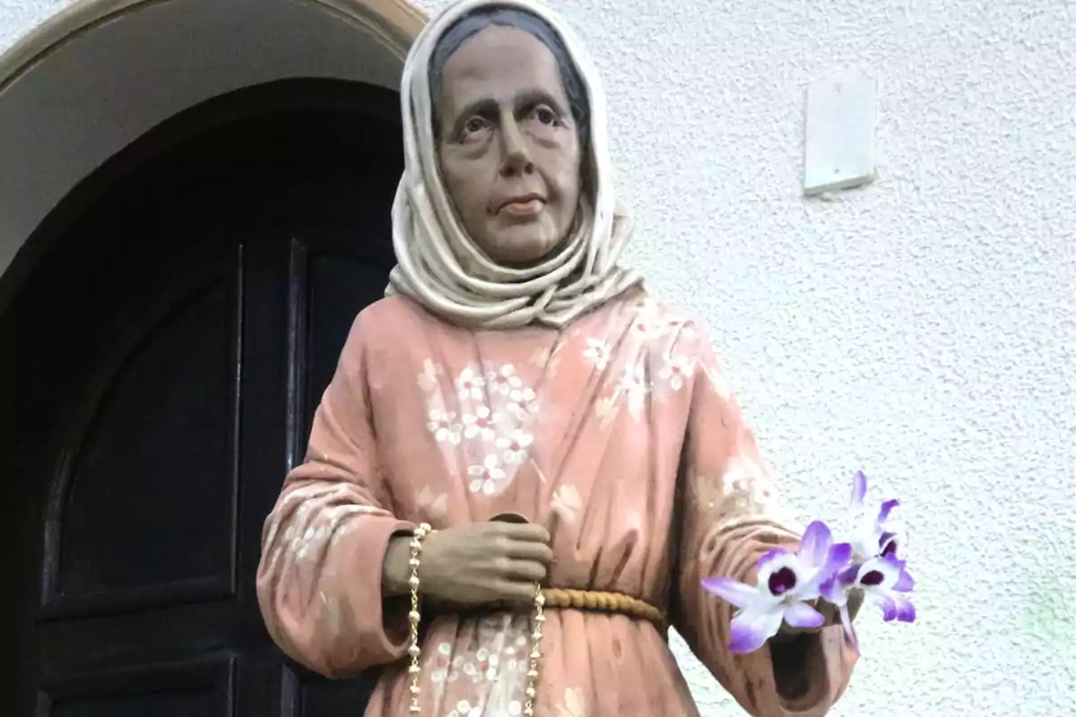 Escultura de una mujer con vestido rosa y pañuelo en la cabeza sosteniendo un rosario y unas flores moradas frente a una puerta oscura Escultura de una mujer con vestido rosa y pañuelo en la cabeza sosteniendo un rosario y unas flores moradas frente a una puerta oscura