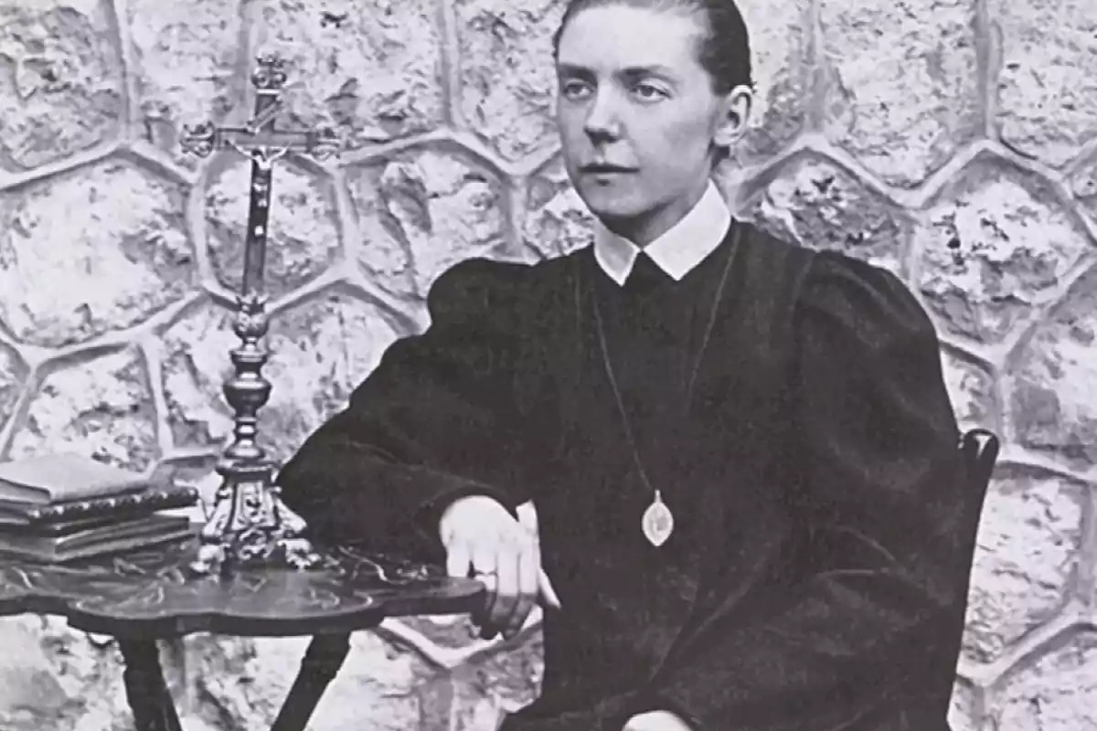 Una mujer sentada junto a una mesa con un crucifijo y libros, con una pared de piedra de fondo. Una mujer sentada junto a una mesa con un crucifijo y libros, con una pared de piedra de fondo.