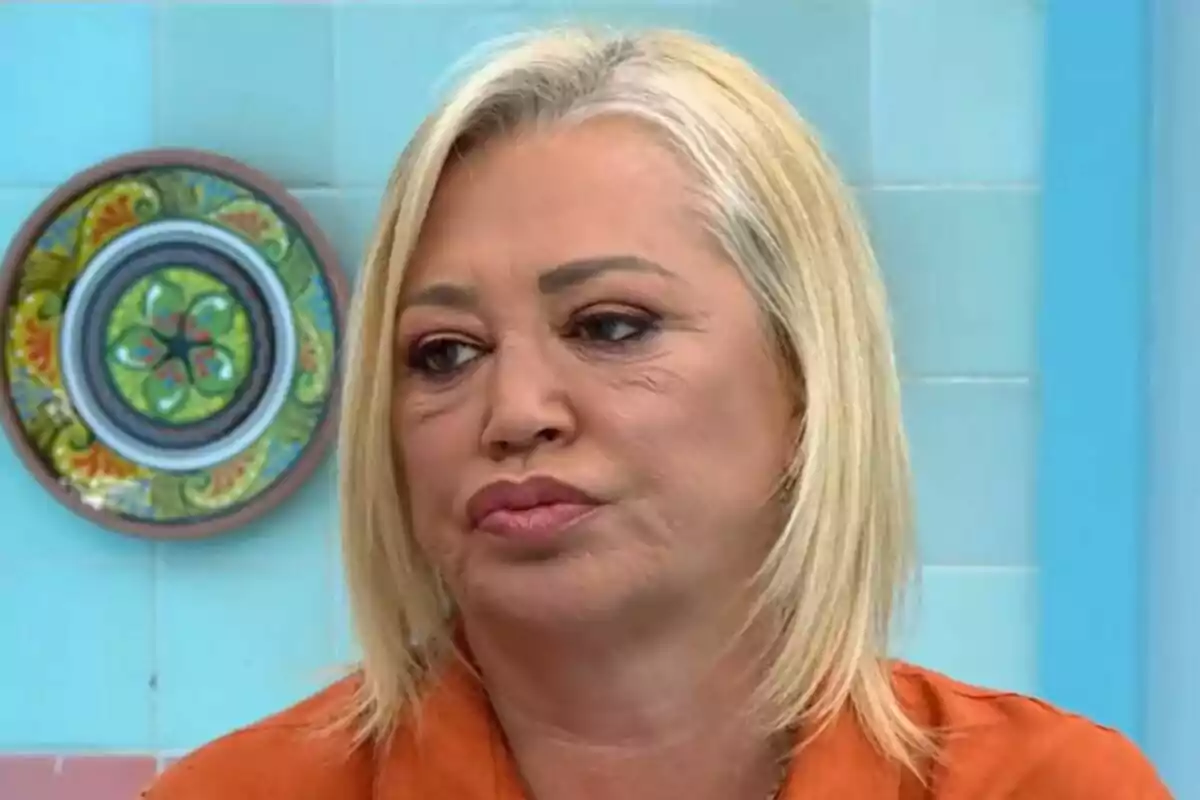 Belén Esteban de cabello rubio y expresión seria en un entorno con azulejos azules y un plato decorativo en la pared en La Familia de la Tele.