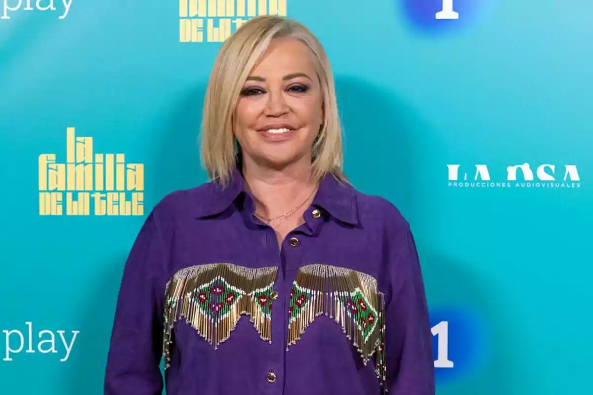 Belén Esteban con cabello rubio y camisa morada decorada, posando frente a un fondo azul con logotipos de Belén Esteban con cabello rubio y camisa morada decorada, posando frente a un fondo azul con logotipos de