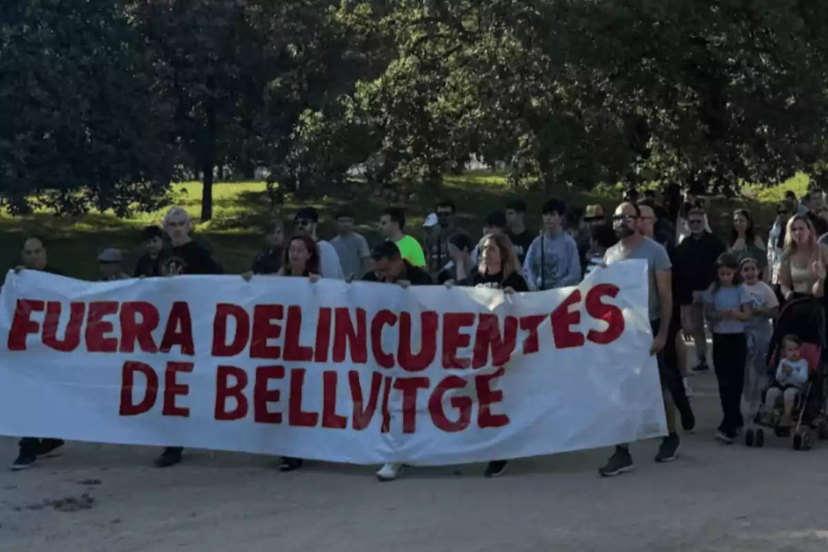 Un grupo de personas sostiene una pancarta que dice "Fuera delincuentes de Bellvitge" mientras caminan por un parque.