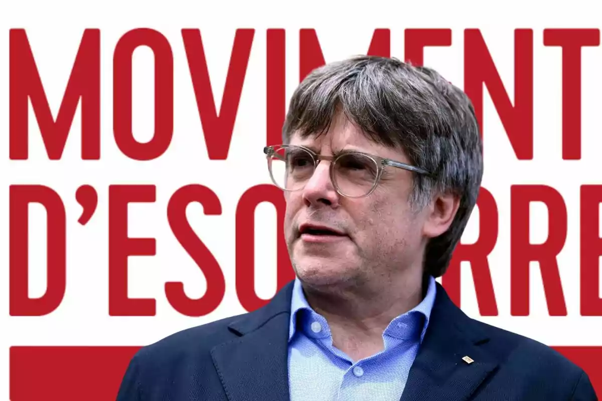 Un hombre con gafas y cabello canoso frente a un cartel rojo con texto en catalán.