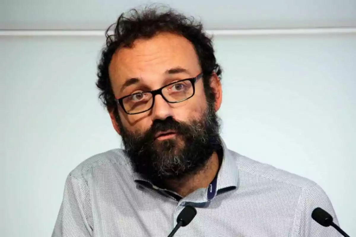 Hombre de barba y gafas hablando frente a dos micrófonos con camisa clara y fondo neutro