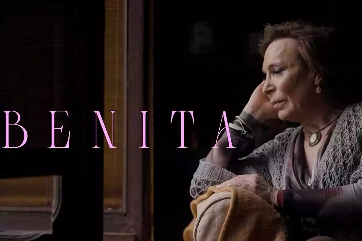 Maestro Joao mayor sentada junto a una ventana mirando pensativa con la palabra BENITA en letras grandes rosas