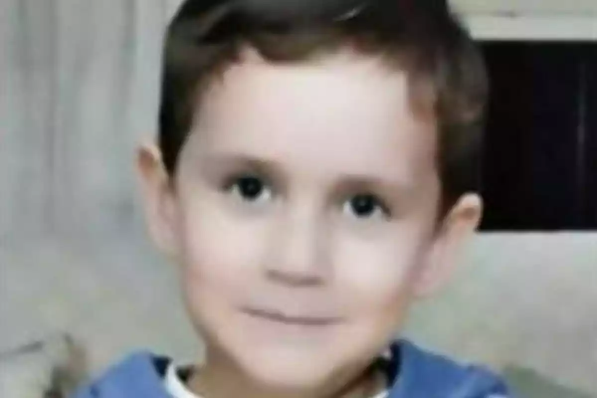 Un niño pequeño con cabello castaño y ojos oscuros, vistiendo una camiseta azul, mira a la cámara con una expresión neutral.