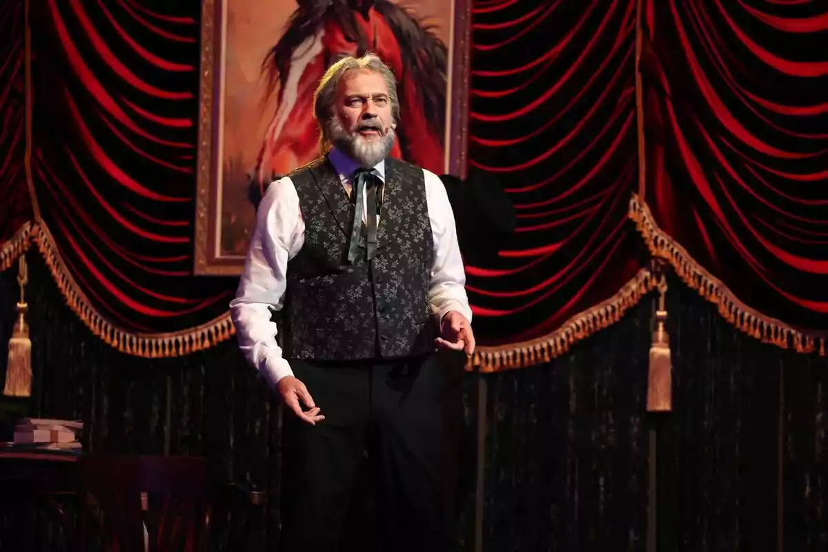 Bertín Osborne con barba y chaleco oscuro está de pie en un escenario de Tu Cara Me Suena decorado con cortinas rojas y un cuadro de un caballo. Bertín Osborne con barba y chaleco oscuro está de pie en un escenario de Tu Cara Me Suena decorado con cortinas rojas y un cuadro de un caballo.