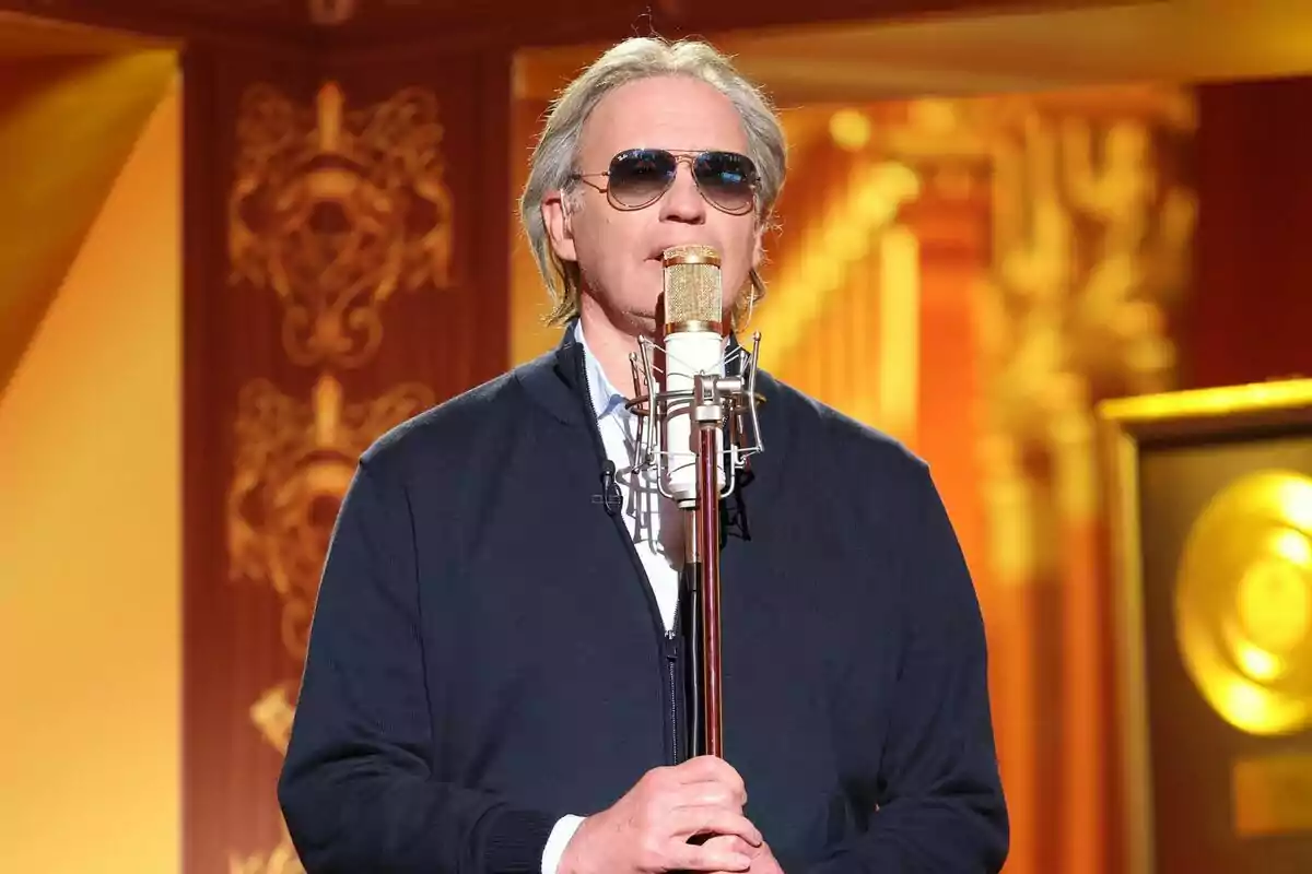 Bertín Osborne de cabello canoso y gafas de sol cantando frente a un micrófono dorado en un escenario decorado elegantemente en Tu Cara Me Suena Bertín Osborne de cabello canoso y gafas de sol cantando frente a un micrófono dorado en un escenario decorado elegantemente en Tu Cara Me Suena