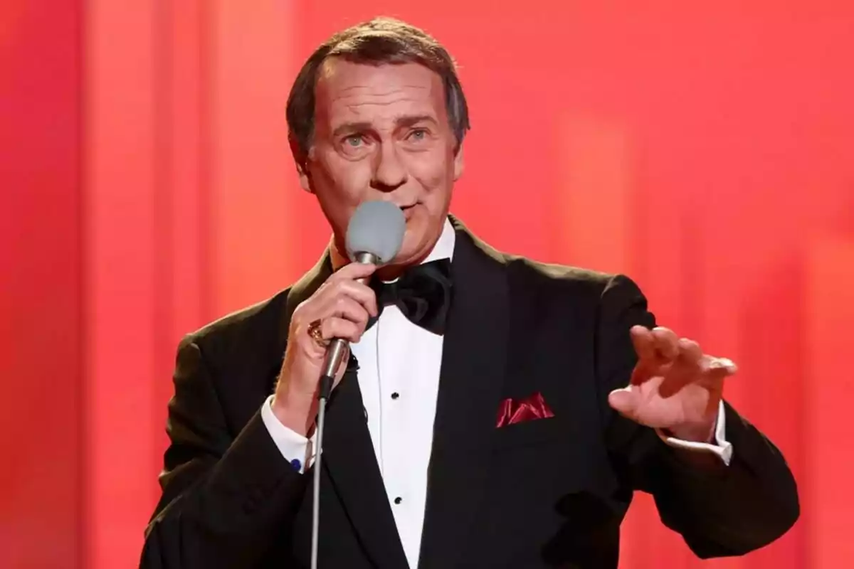 Bertín Osborne en un escenario con un micrófono en la mano vestido de esmoquin negro con pajarita y pañuelo rojo en el bolsillo del saco fondo rojo en Tu Cara Me Suena. Bertín Osborne en un escenario con un micrófono en la mano vestido de esmoquin negro con pajarita y pañuelo rojo en el bolsillo del saco fondo rojo en Tu Cara Me Suena.