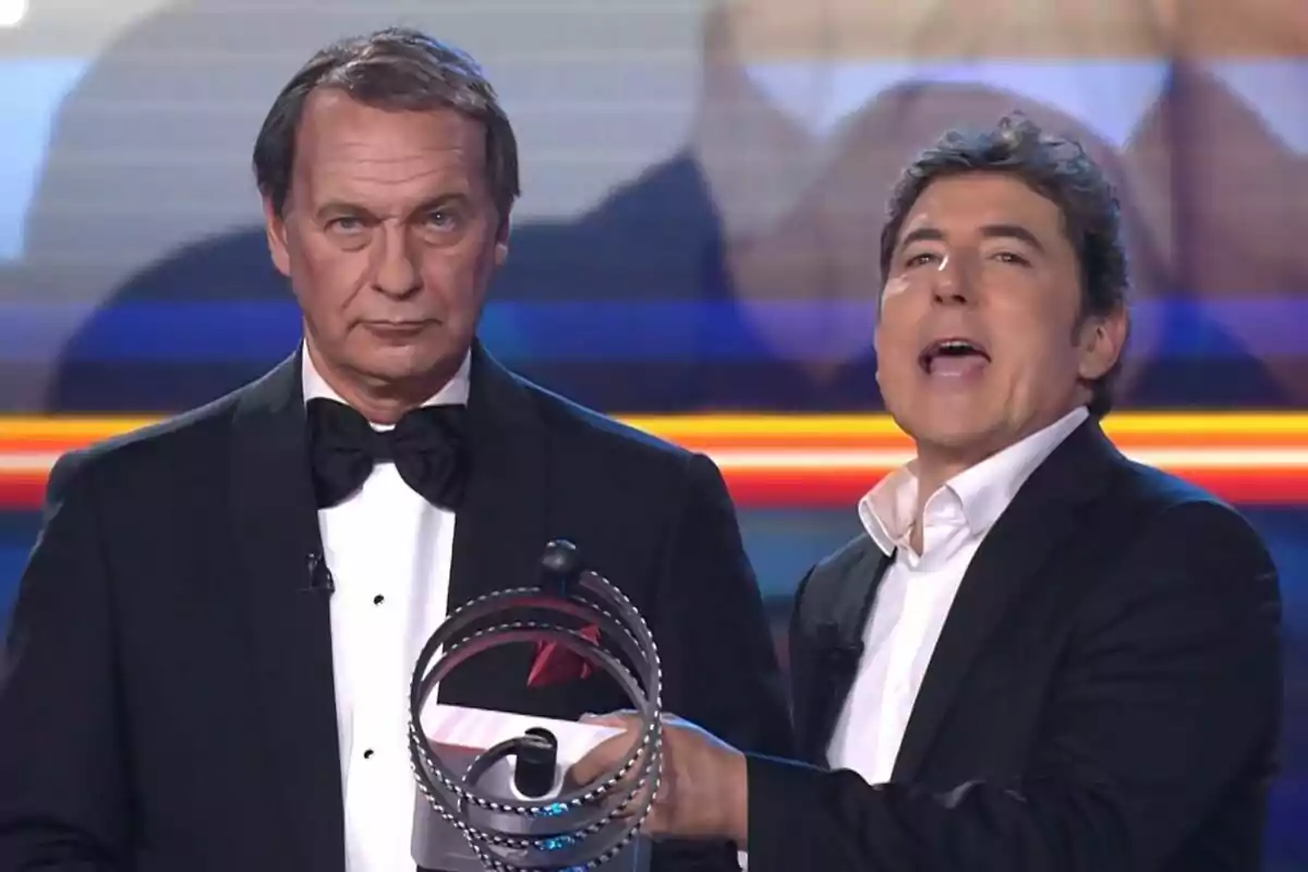 Dos hombres en un escenario, Manel Fuentes sostiene un objeto circular mientras Bertín Osborne lleva un esmoquin y corbata de moño en Tu Cara Me Suena.