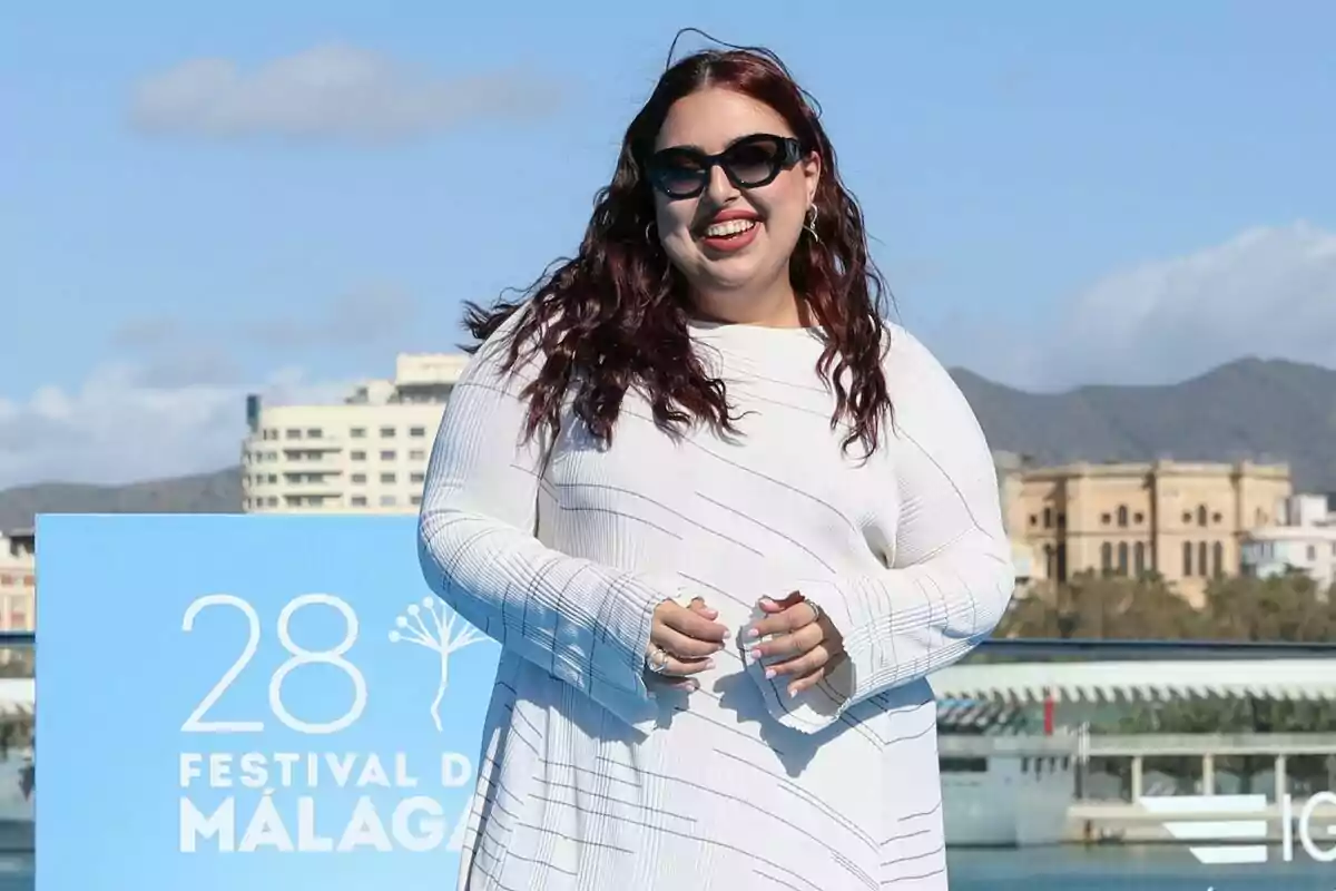 Blanca Martínez con gafas de sol y cabello largo sonríe frente a un cartel del Festival de Málaga con un edificio y montañas al fondo en la presentación de Mariliendre. Blanca Martínez con gafas de sol y cabello largo sonríe frente a un cartel del Festival de Málaga con un edificio y montañas al fondo en la presentación de Mariliendre.