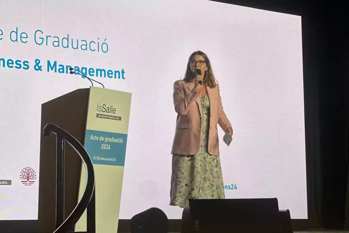 Una mujer con un micrófono en la mano está de pie junto a un podio en un evento de graduación de la Universidad Ramon Llull, La Salle, para la promoción de 2024.