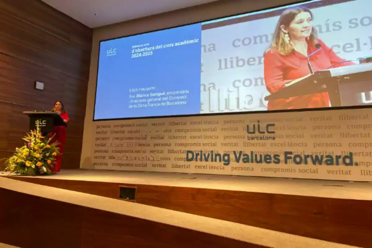 Blanca Sorigué en un podio dando un discurso en un evento académico en un auditorio con una pantalla grande que muestra su imagen y texto relacionado con la inauguración del curso académico 2024-2025 en la ULC.