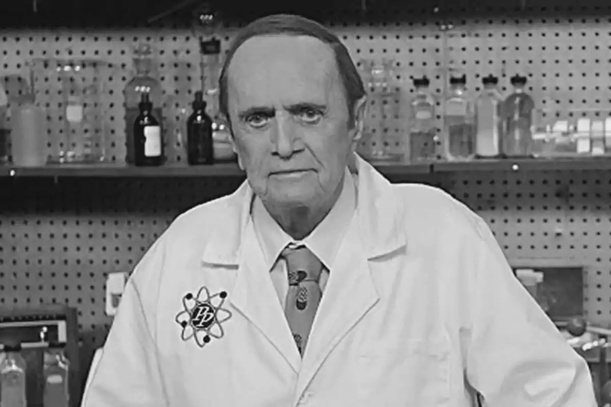 Fotografía en blanco y negro de Bob Newhart con bata de laboratorio y corbata, de pie en un laboratorio con estantes llenos de frascos y botellas detrás de él en la serie The Big Bang Theory