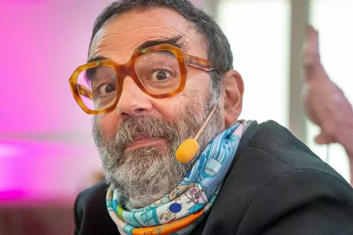 Un hombre con barba y gafas grandes lleva un micrófono de diadema y una bufanda colorida.