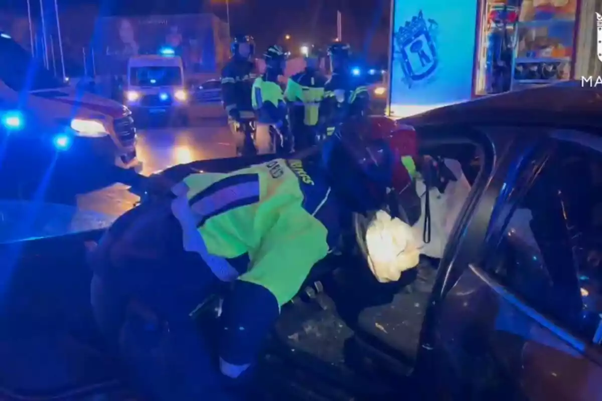 Un grupo de bomberos y personal de emergencia trabaja en la escena de un accidente de tráfico durante la noche, con luces azules de vehículos de emergencia iluminando el área.