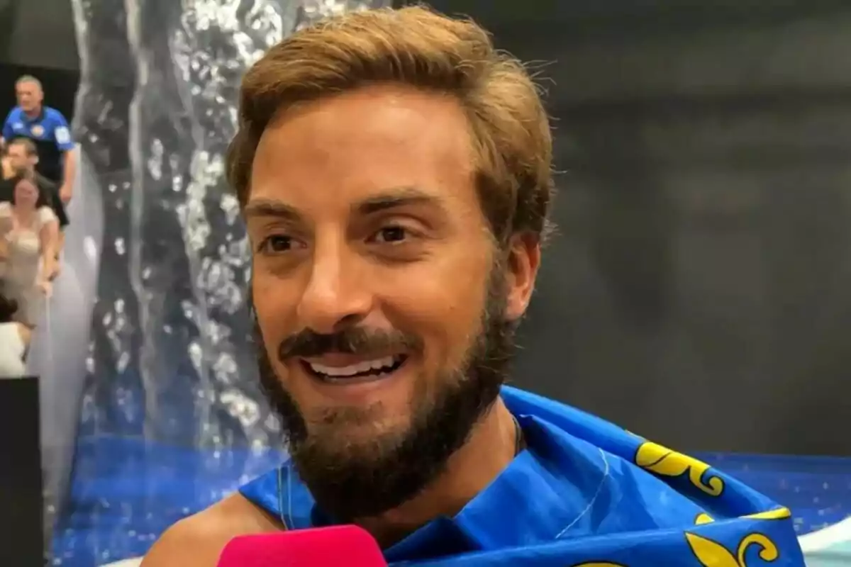 Borja González sonriente con barba y cabello claro envuelto en una bandera de la Comunitat Valenciana azul con detalles amarillos mientras sostiene un micrófono rosa en el plató de Supervivientes