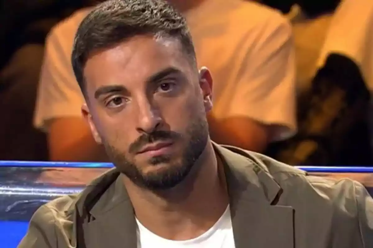 Borja González con barba y cabello corto mira hacia adelante con expresión seria, vistiendo una chaqueta marrón claro y una camiseta blanca, en el plató de La Isla de las Tentaciones.