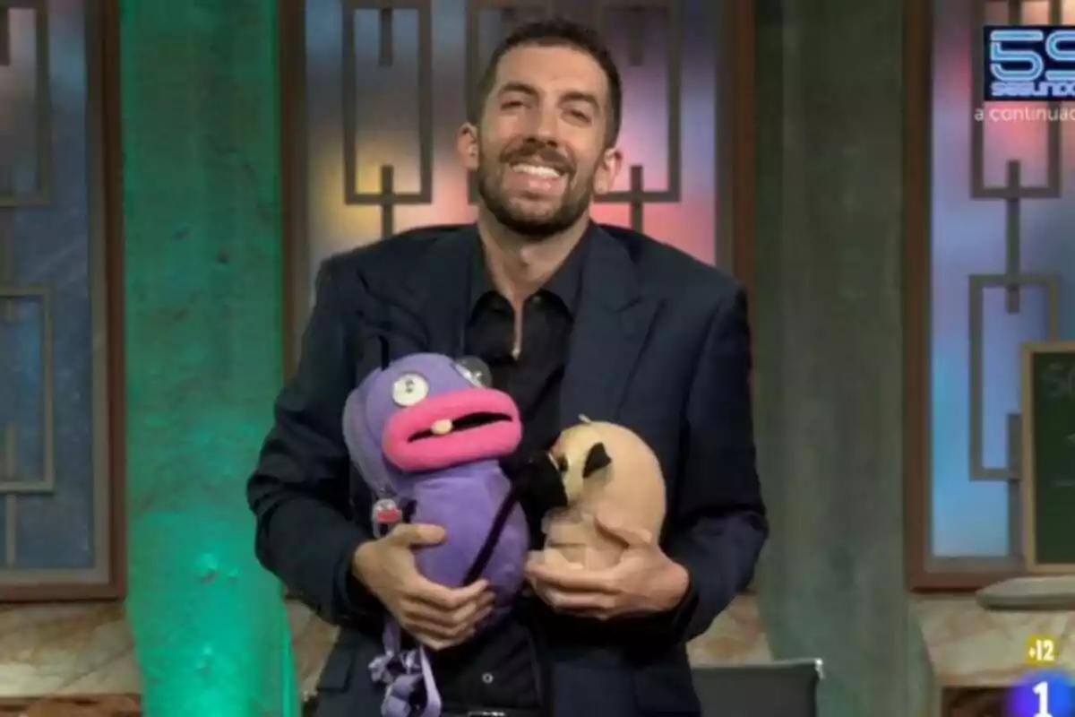 David Broncano sonriente en La Revuelta sostiene dos marionetas, una morada con labios rosados que es una hormiga de EL Hormiguero y otra beige con orejas negras, en un set de televisión con fondo verde y elementos decorativos geométricos.