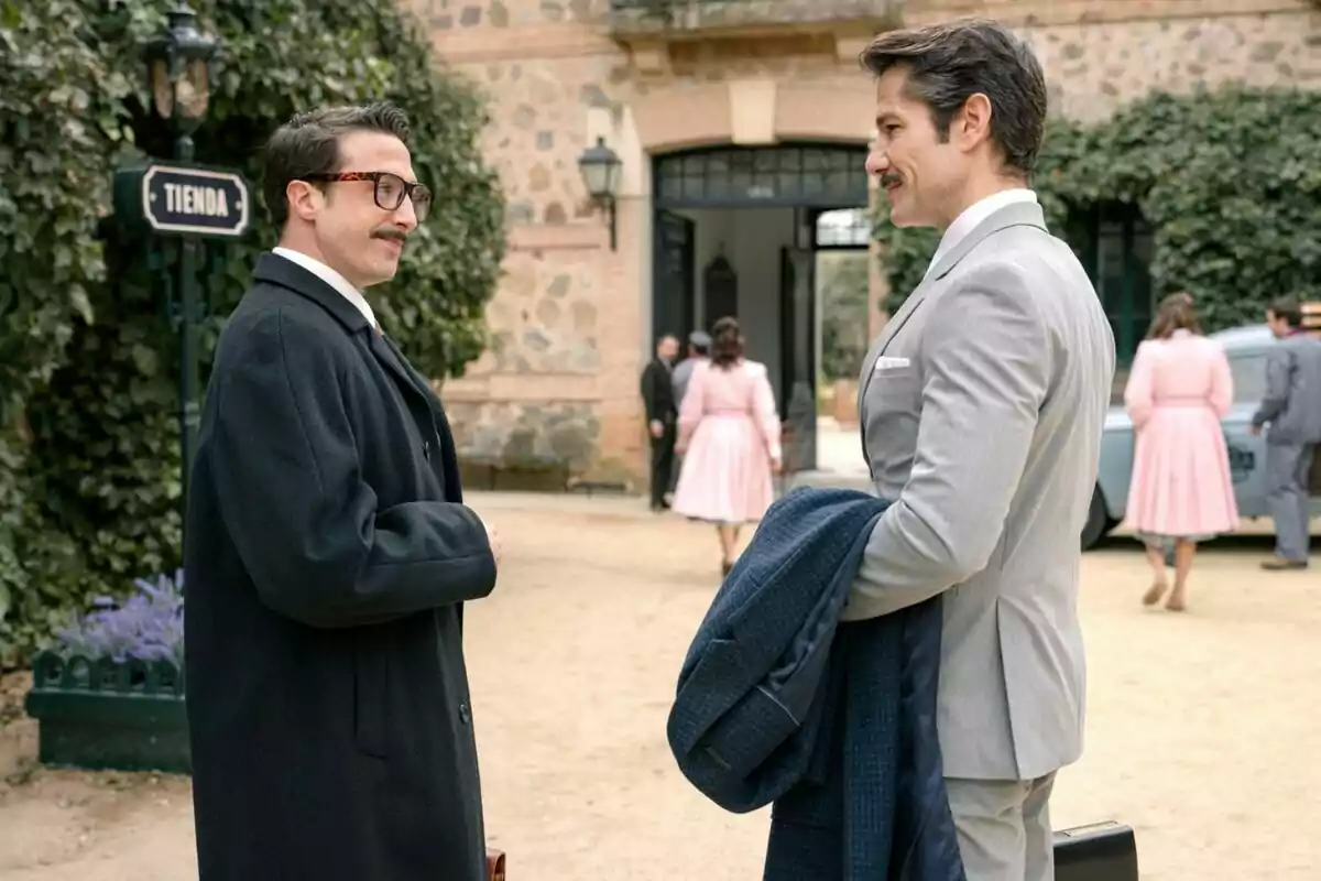 Bruno Tamarit y Alejandro Albarracín elegantemente vestidos conversan al aire libre frente a un edificio antiguo mientras varias personas caminan en el fondo en Sueños de Libertad Bruno Tamarit y Alejandro Albarracín elegantemente vestidos conversan al aire libre frente a un edificio antiguo mientras varias personas caminan en el fondo en Sueños de Libertad