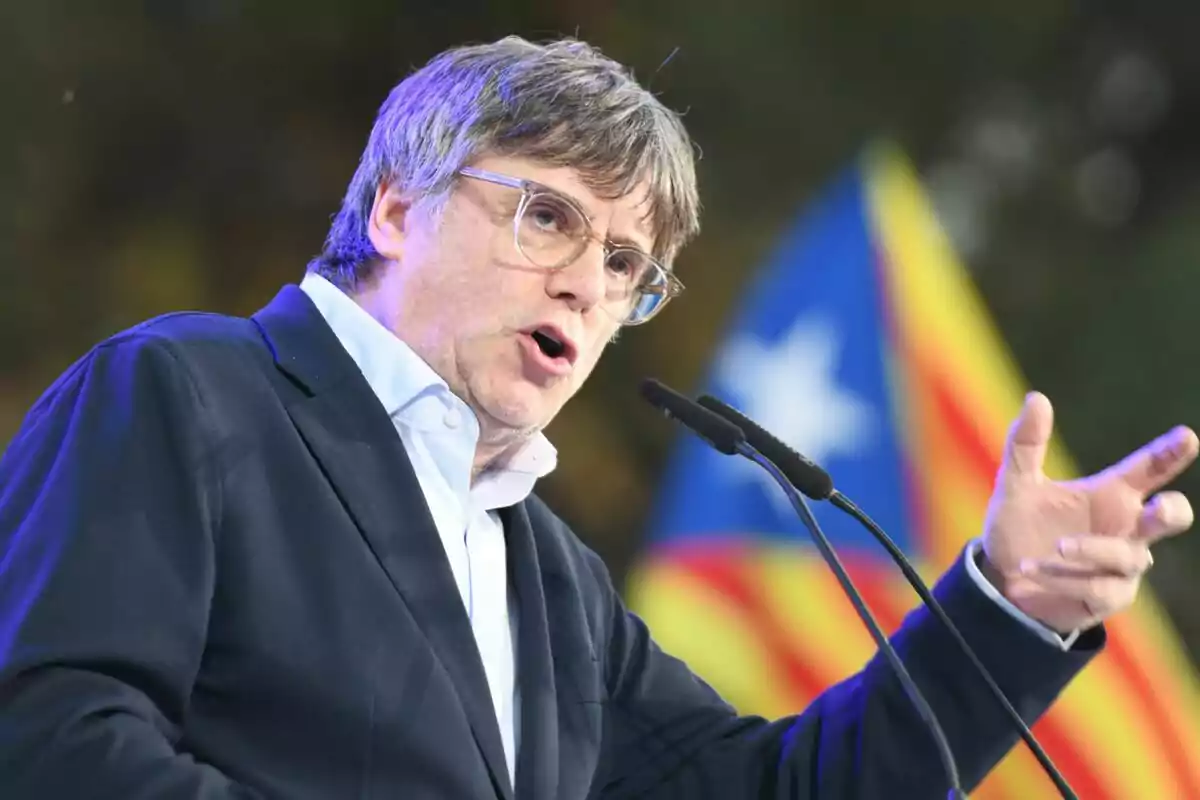 Carles Puigdemont hablando en un micrófono con una bandera catalana de fondo.