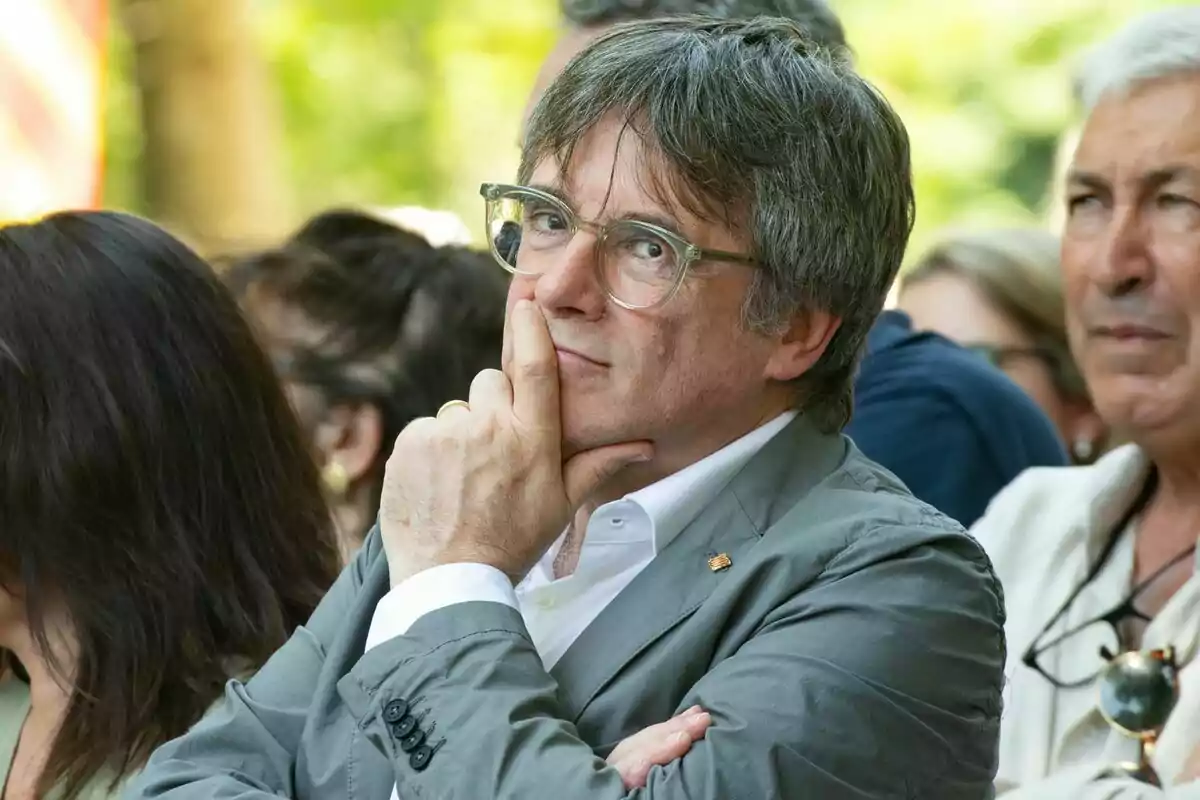 Carles Puigdemont con chaqueta gris, con la mano en la barbilla, parece pensativo en un evento al aire libre.