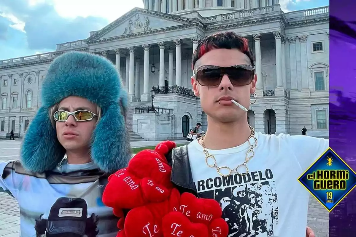 Ca7riel y Paco Amoroso posan frente a un edificio con columnas, una lleva un gorro azul y gafas de sol, la otra tiene gafas oscuras y un collar dorado, sostienen un ramo de corazones rojos con la frase "Te amo", junto a el logo de El Hormiguero