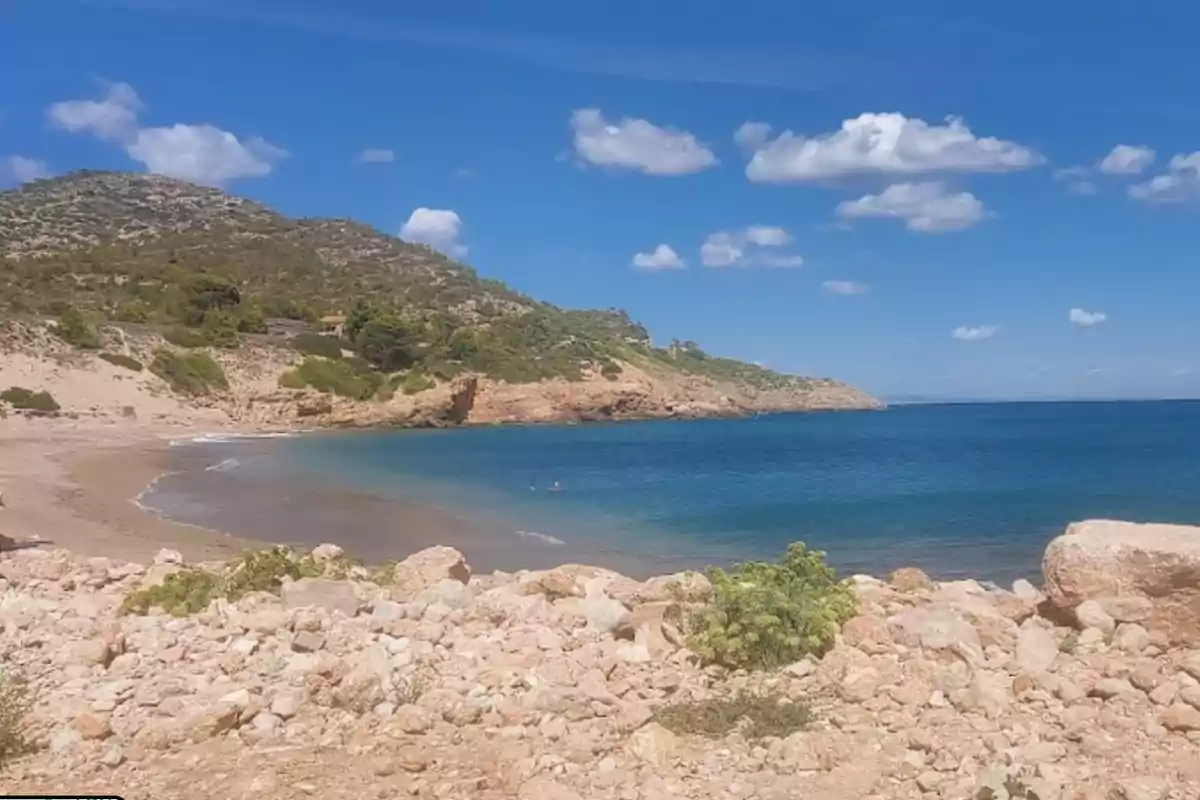 Playa con aguas cristalinas, arena y rocas, rodeada de colinas verdes bajo un cielo azul con nubes dispersas. Playa con aguas cristalinas, arena y rocas, rodeada de colinas verdes bajo un cielo azul con nubes dispersas.