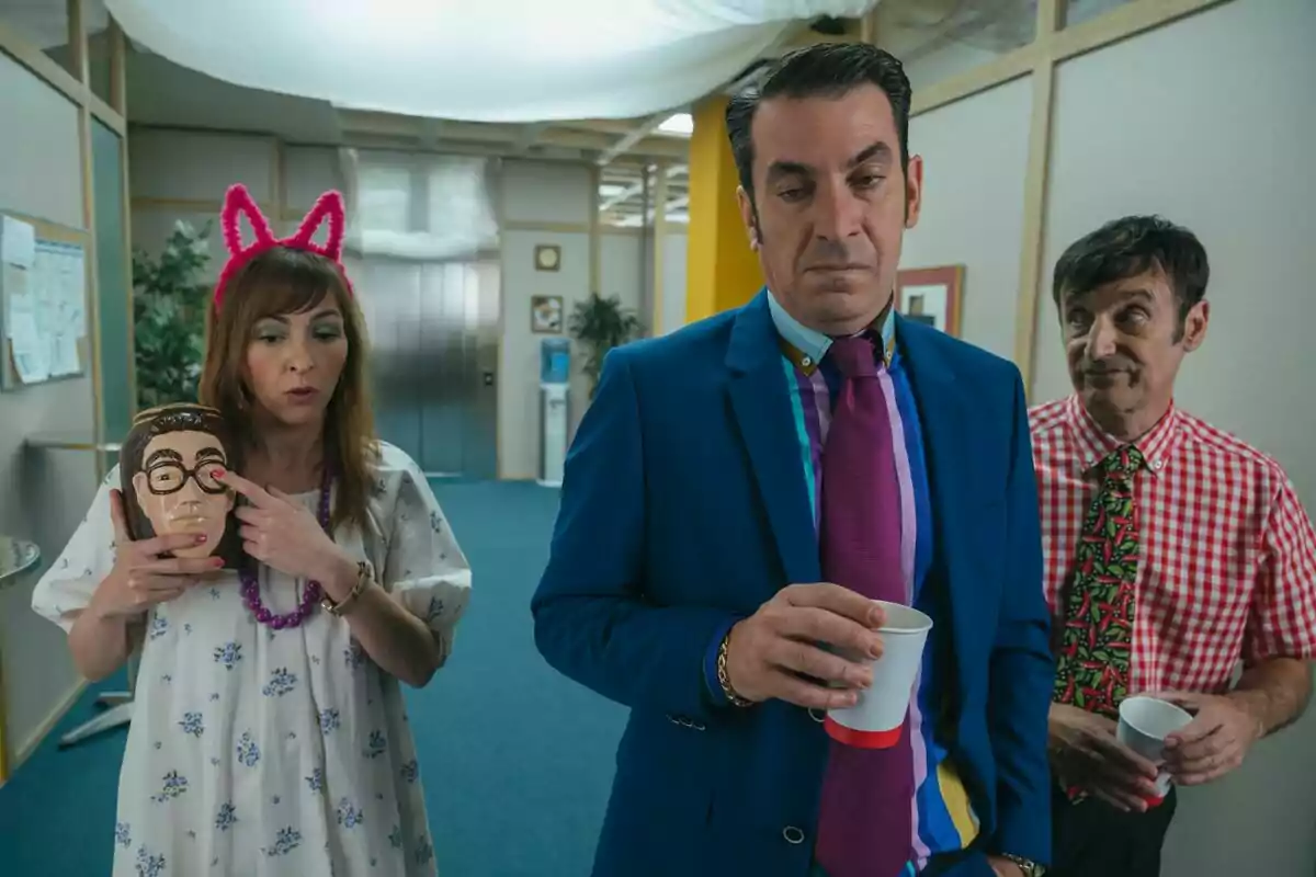 Tres personas en una oficina de la película de Camera Café, una mujer con orejas rosas sostiene una máscara, Arturo Valls de traje azul y corbata morada sostiene un vaso y Carlos Chamarro con camisa de cuadros y corbata colorida los observa