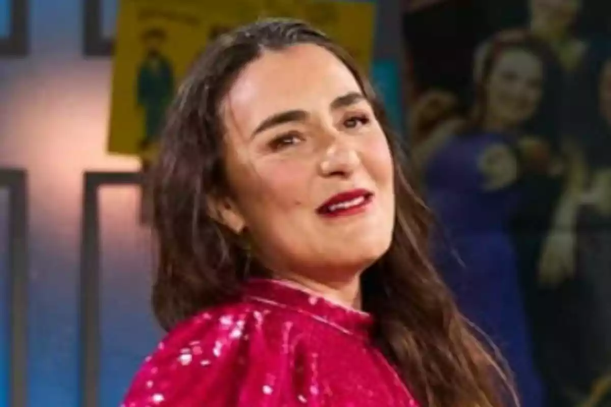 Candela Peña con cabello largo y oscuro lleva un vestido rojo brillante y está sonriendo ligeramente mientras mira hacia un lado en un entorno interior con luces y carteles de fondo en 'La Revuelta'.