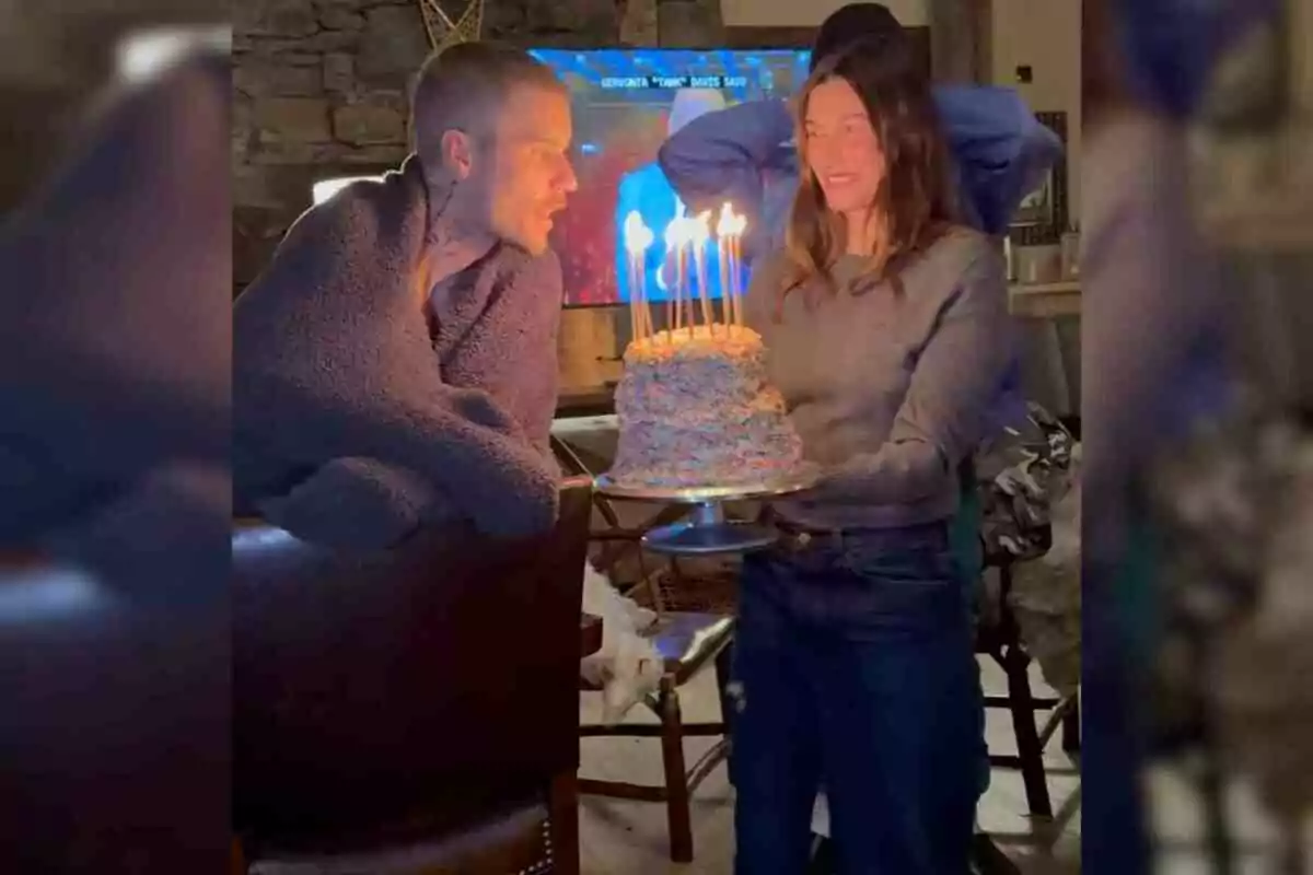 Justin Bieber envuelta en una manta observa un pastel de cumpleaños con velas encendidas que sostiene Hailey Bieber sonriente. Justin Bieber envuelta en una manta observa un pastel de cumpleaños con velas encendidas que sostiene Hailey Bieber sonriente.