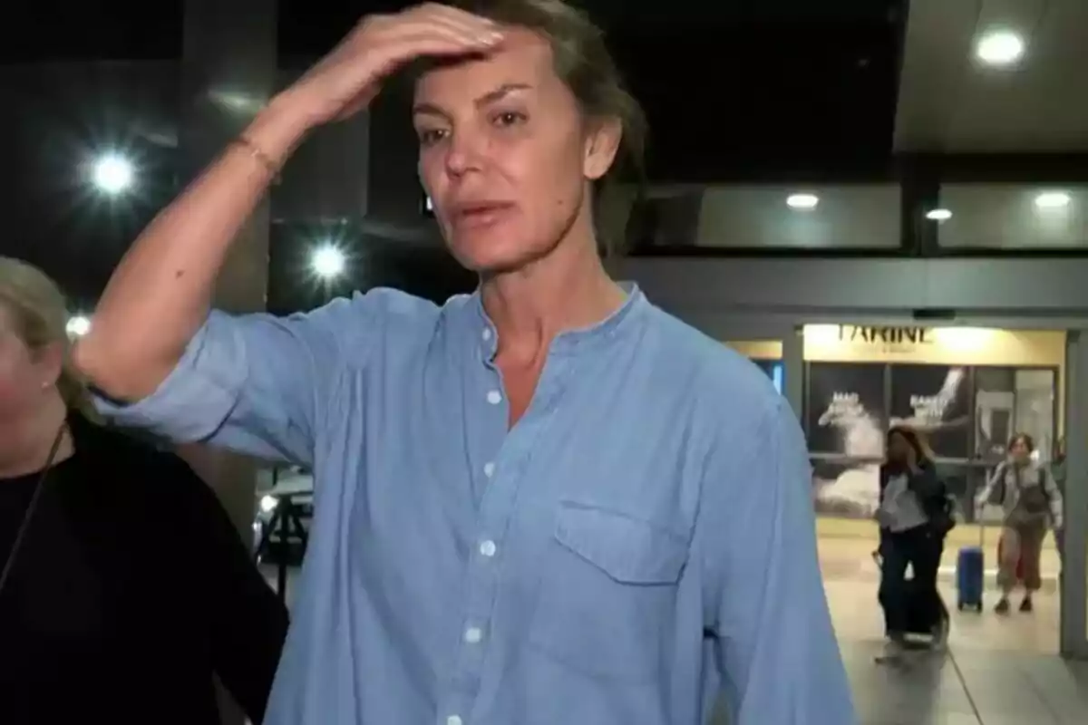 Una mujer con camisa azul claro se toca la frente mientras está de pie en un lugar público iluminado. Una mujer con camisa azul claro se toca la frente mientras está de pie en un lugar público iluminado.
