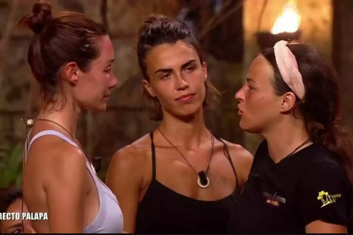 Tres mujeres conversan intensamente en un entorno al aire libre, con una fogata de fondo y la palabra 