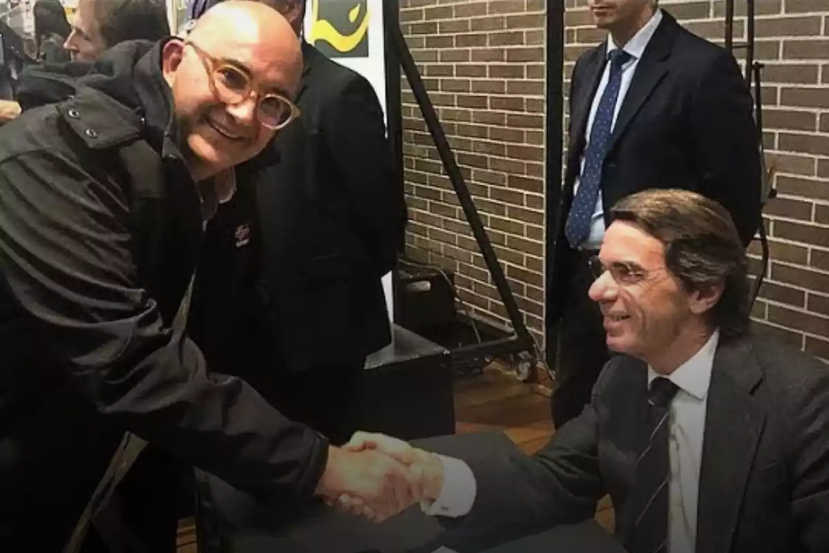 Al lado izquierdo, Xavier Rius un poco agachado, sonriendo y dándole la mano a José María Aznar sentado y sonriendo