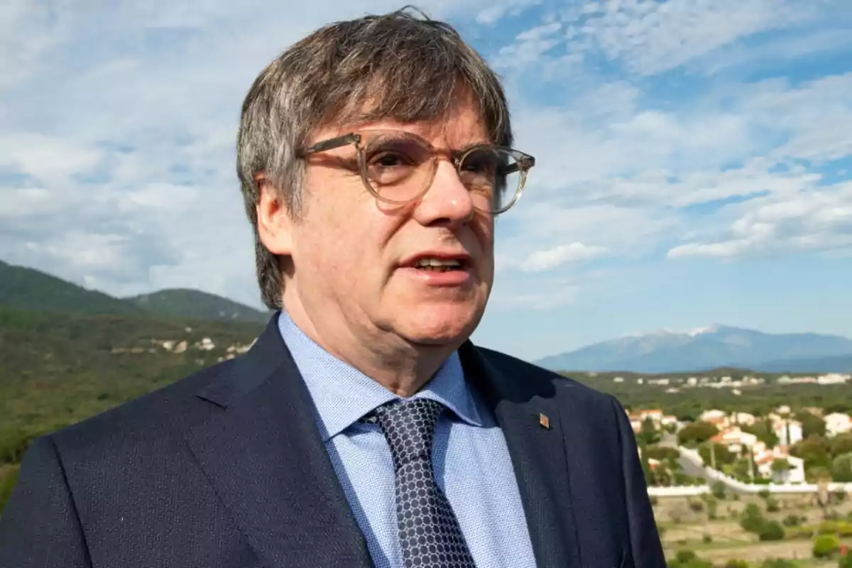 Hombre con gafas y traje oscuro en un entorno natural con montañas al fondo.