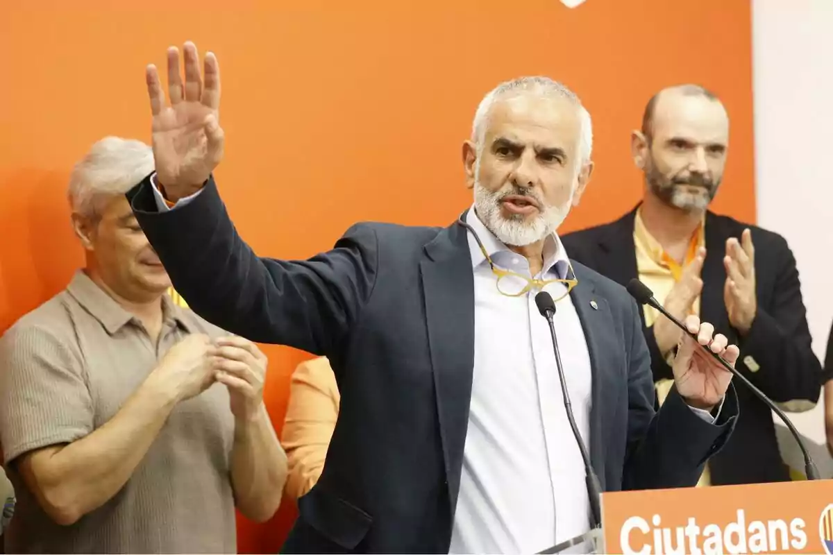 Un hombre de cabello canoso y barba está hablando en un micrófono con la mano levantada mientras otras personas aplauden en el fondo sobre un fondo naranja.