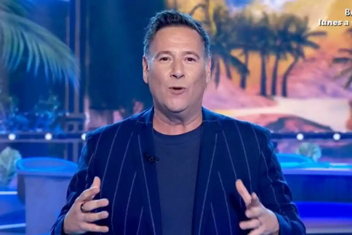 Carlos Latre con una chaqueta de rayas habla frente a un fondo colorido con palmeras en Babylon Show