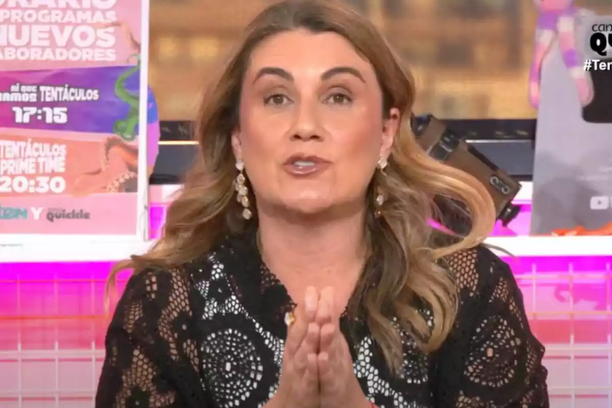 Carlota Corredera con cabello largo y suelto, vestida con una blusa negra de encaje, está hablando frente a la cámara en 'Tentáculos', con carteles coloridos en el fondo.