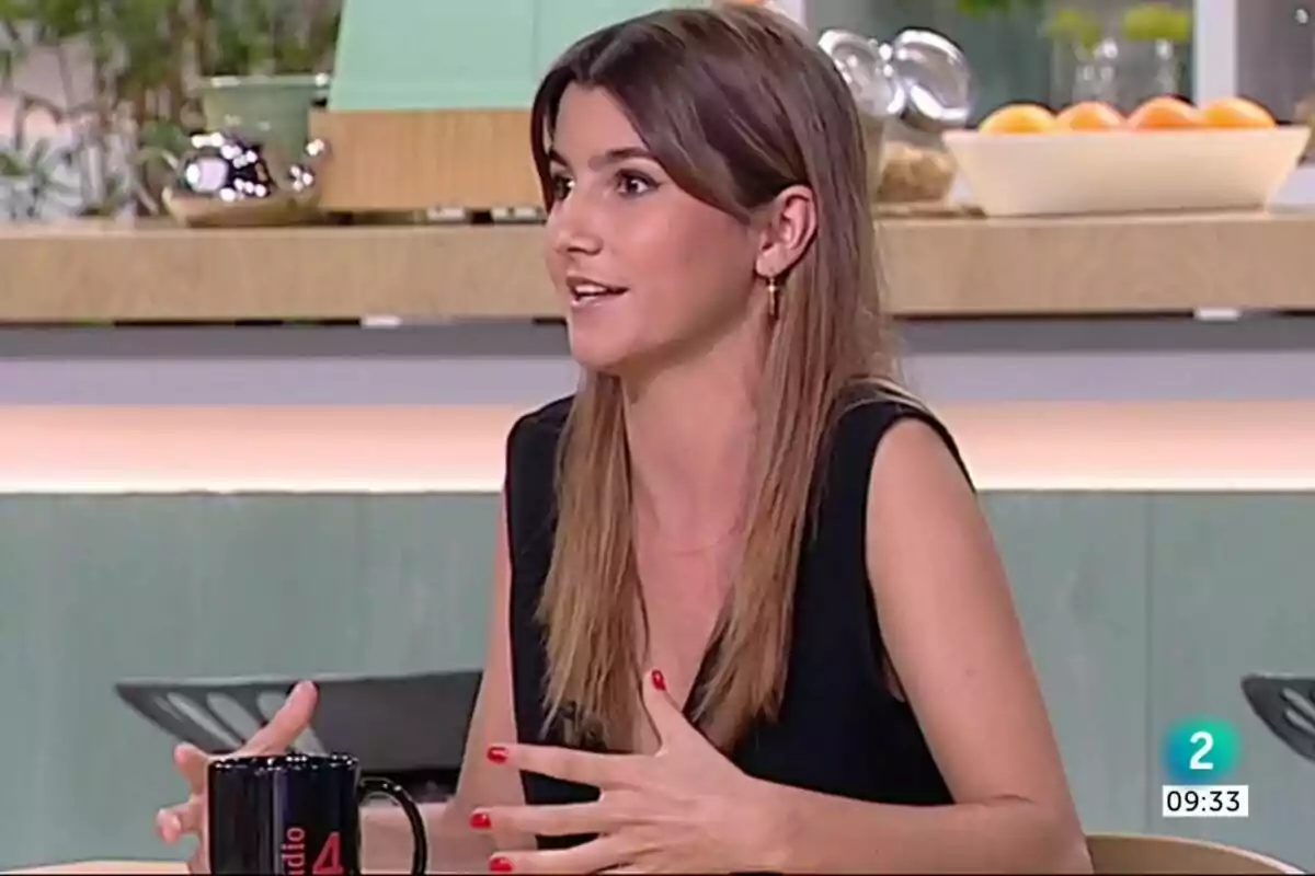 Una mujer con cabello largo y castaño está sentada en una mesa, hablando y gesticulando con las manos; lleva una blusa negra sin mangas y en la mesa hay una taza negra con el logo de Una mujer con cabello largo y castaño está sentada en una mesa, hablando y gesticulando con las manos; lleva una blusa negra sin mangas y en la mesa hay una taza negra con el logo de