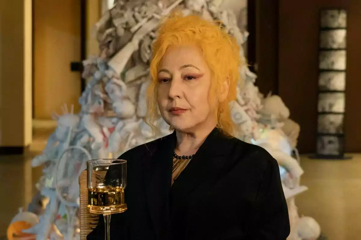 Carmen Machi con cabello amarillo sosteniendo un vaso de bebida, con una escultura abstracta en el fondo en Furia.