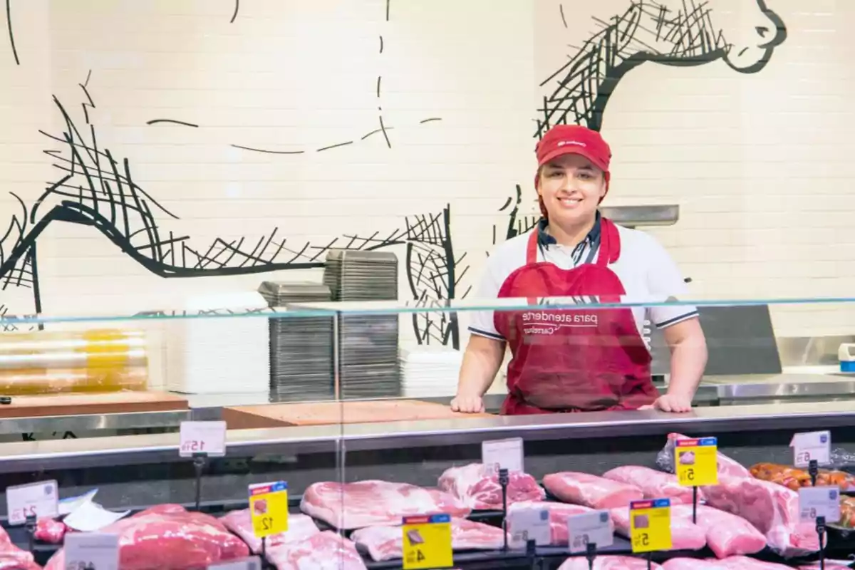 Persona con delantal rojo y gorra roja atendiendo en una carnicería con varios cortes de carne en exhibición.