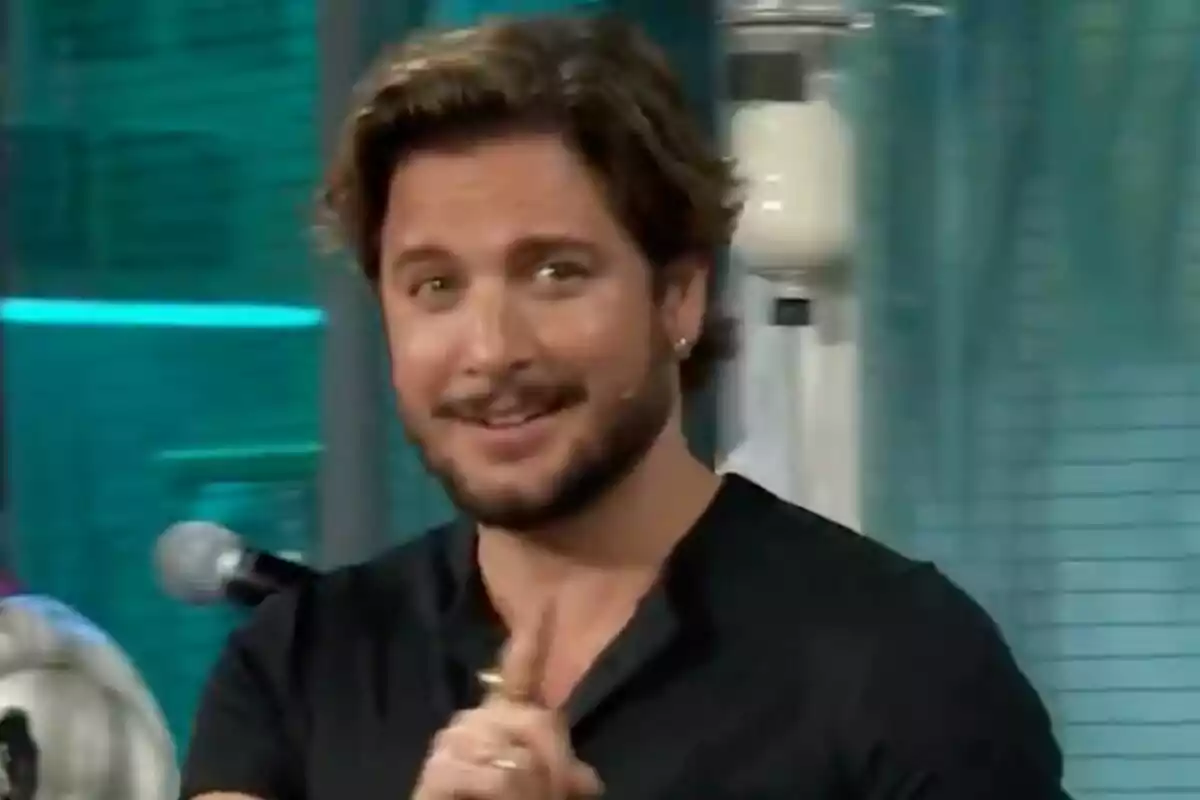 Manuel Carrasco con barba y cabello castaño sonríe mientras señala con el dedo en un entorno interior con luces azules en 'La Revuelta'.