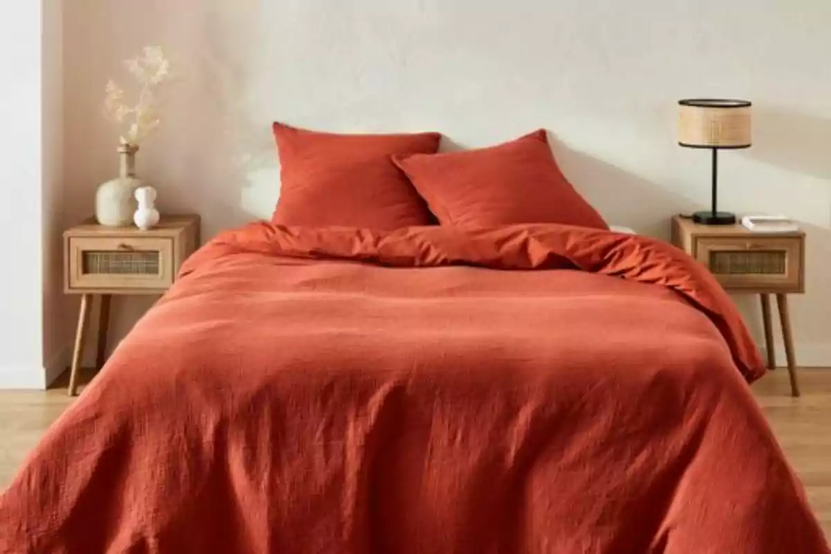 Una cama con edredón y almohadas de color terracota, flanqueada por dos mesitas de noche de madera, una con una lámpara y la otra con un jarrón decorativo. Una cama con edredón y almohadas de color terracota, flanqueada por dos mesitas de noche de madera, una con una lámpara y la otra con un jarrón decorativo.